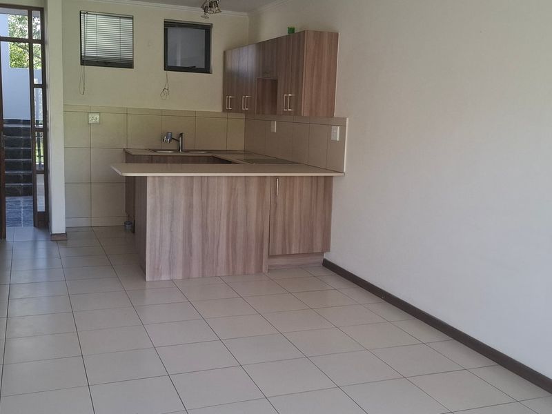 An open-plan living and kitchen area featuring neutral ceramic floor tiles, laminate cabinetry, and a functional breakfast counter in a clean, vacant space.