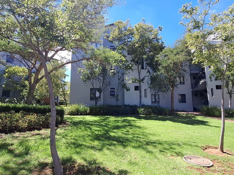 A view of the apartment complex exterior featuring a well-maintained garden, mature trees, and a covered parking area.