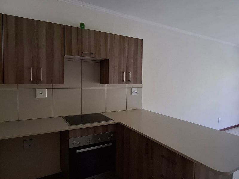 Open-plan kitchen featuring wood-look cabinetry, built-in oven and hob, and a breakfast bar, flowing into a bright living area with access to a balcony.