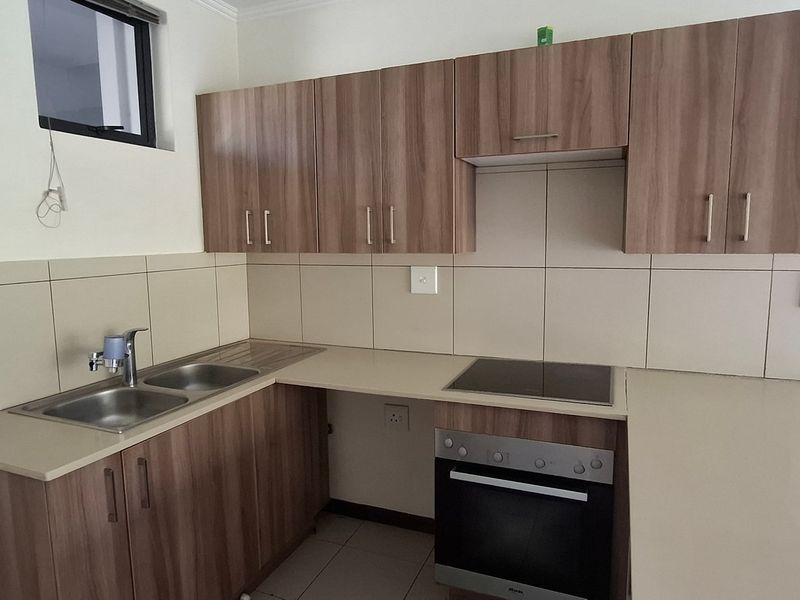 A clean, functional kitchen featuring wood-finish cabinetry, stone-look countertops, a built-in electric oven, and a stainless steel double sink with tiled splashbacks.