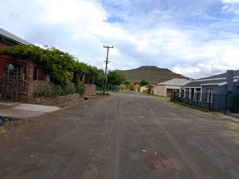 A street view showing the neighbourhood approach with residential properties, mature landscaping, and a scenic mountain backdrop.