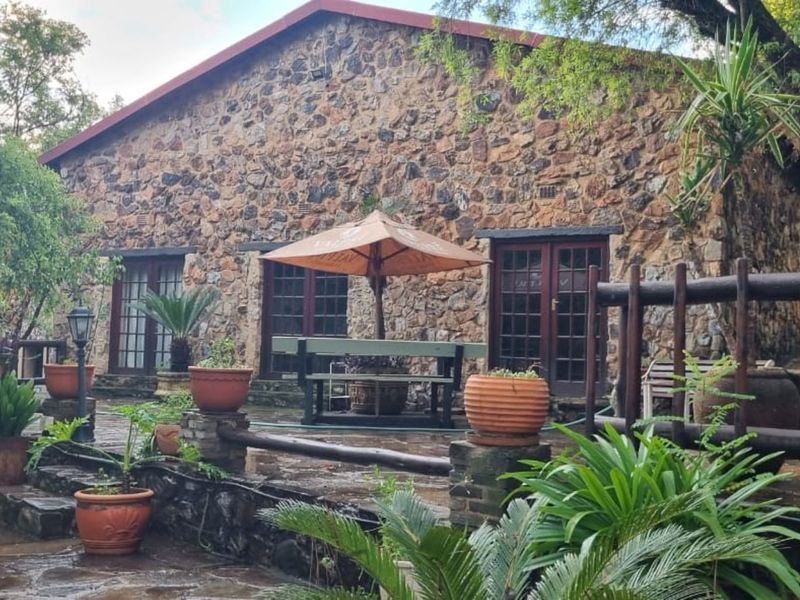 A rustic stone-clad farmhouse exterior featuring a paved patio area, timber-framed doors, and a natural stone retaining wall set within a lush garden.