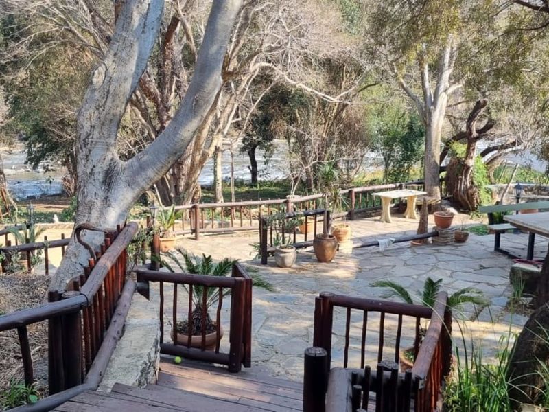A rustic outdoor patio area featuring stone paving and timber railings, overlooking a natural wooded landscape and river setting.