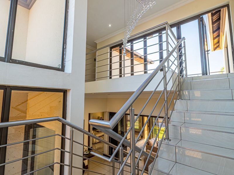 A bright, modern double-volume hallway featuring a tiled staircase with a sleek stainless steel balustrade and expansive windows.