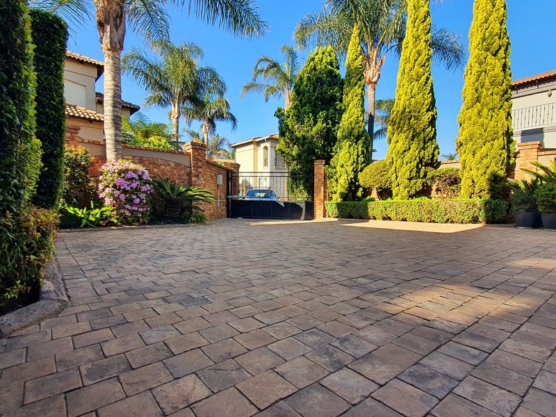 A well-maintained paved driveway leading to a secure gated entrance, featuring established landscaping, brick boundary walls, and mature trees.