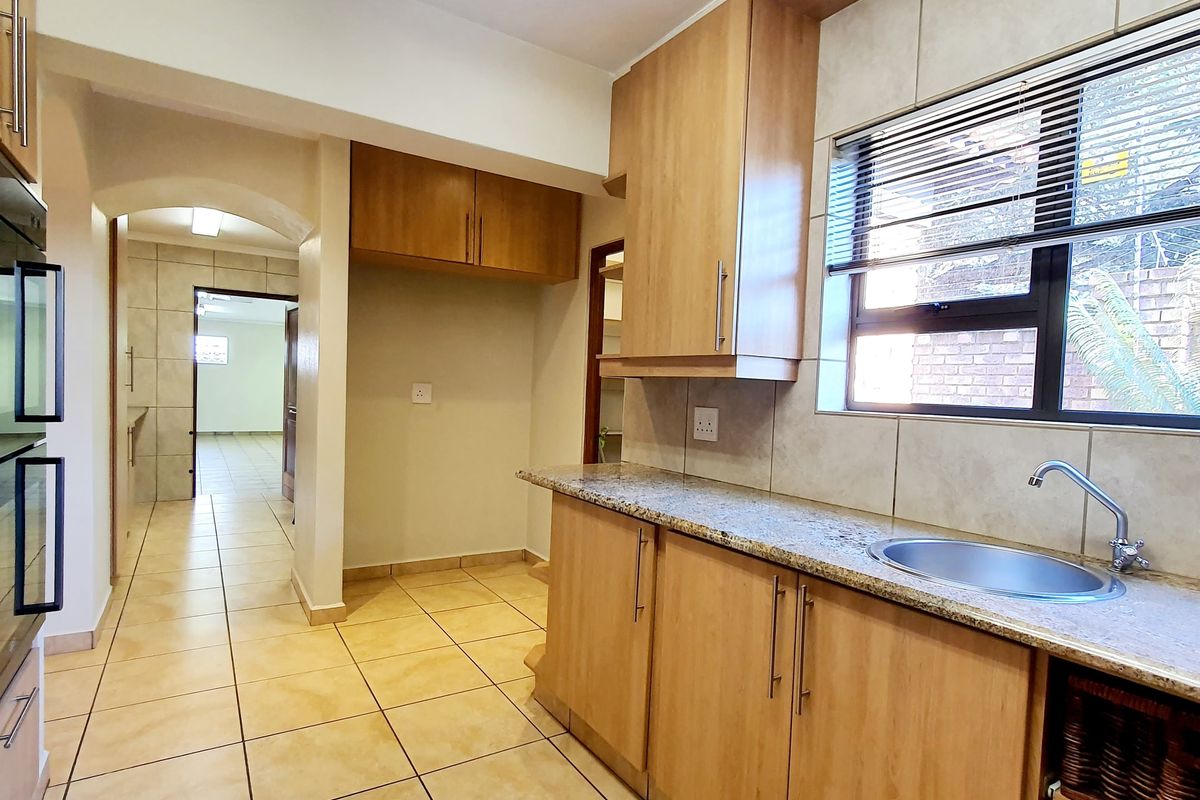 A functional kitchen featuring light wood cabinetry, granite countertops, a stainless steel sink, and tiled flooring, leading into an adjacent hallway.
