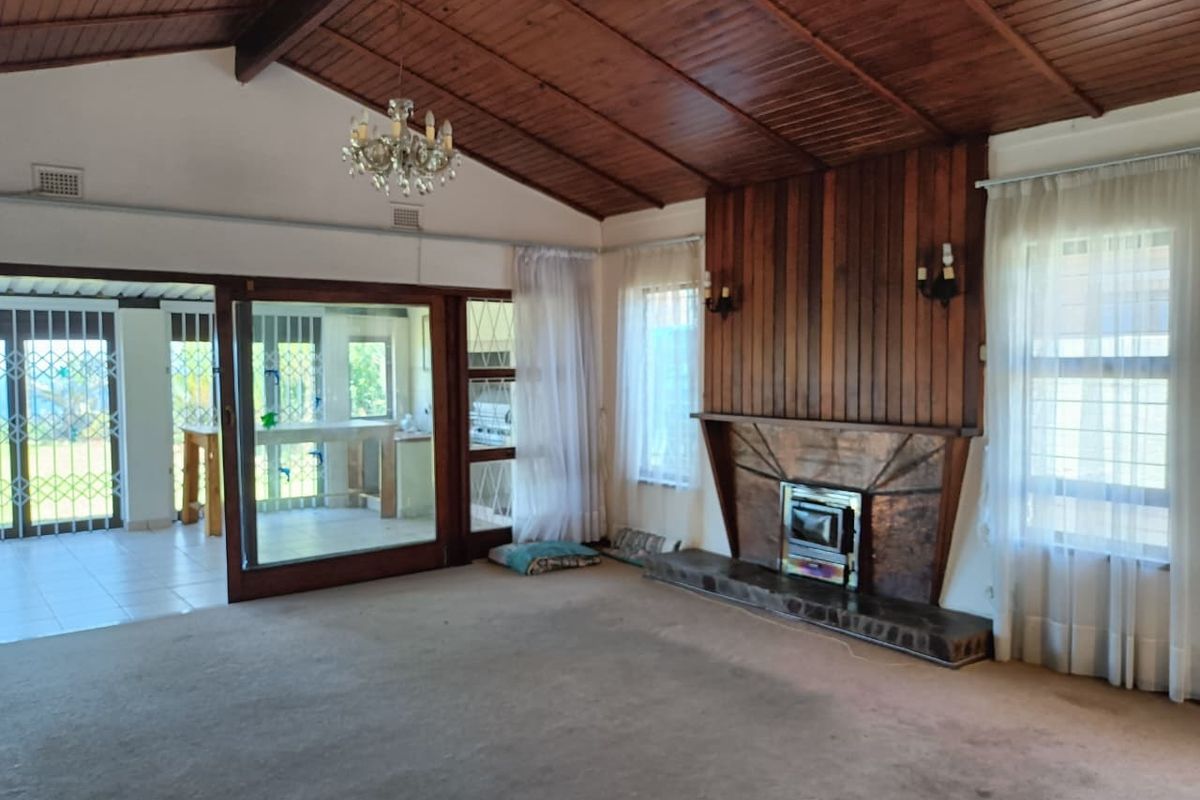 Spacious living room featuring a vaulted timber ceiling, wood-paneled accent wall, and a built-in fireplace with a stone hearth, opening to an outdoor area.