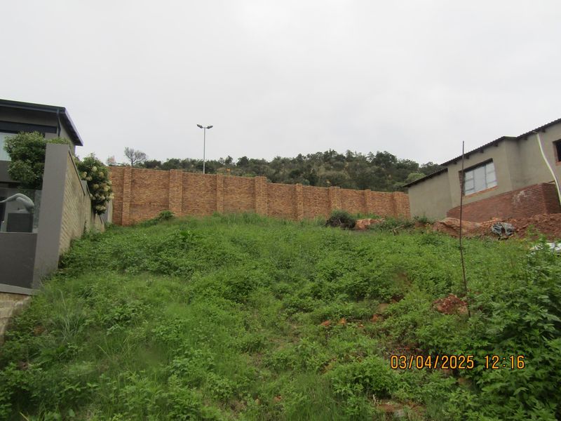 A view of a sloped residential plot featuring a brick boundary wall and surrounding vegetation.