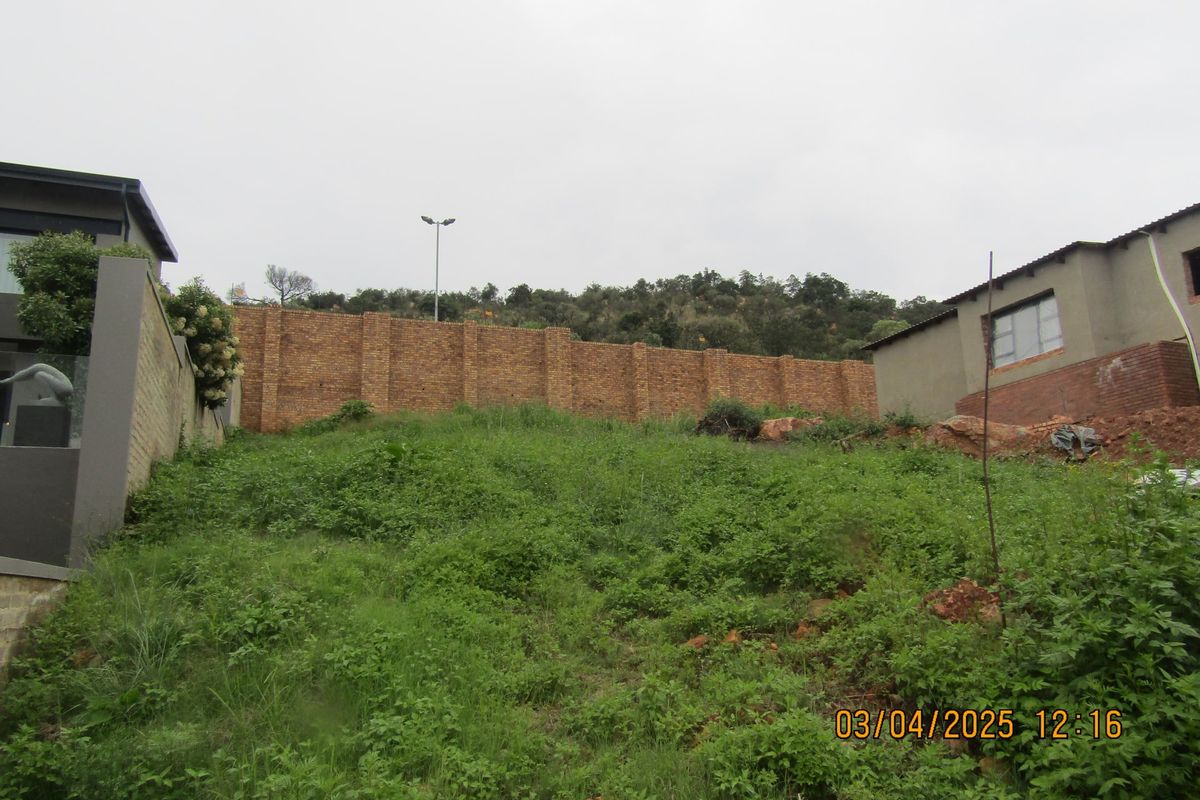 A view of a sloped residential plot featuring a brick boundary wall and surrounding vegetation.