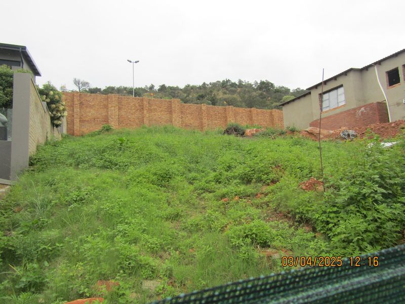 A sloped, undeveloped residential plot featuring a brick boundary wall and overgrown vegetation, situated between existing neighboring properties.