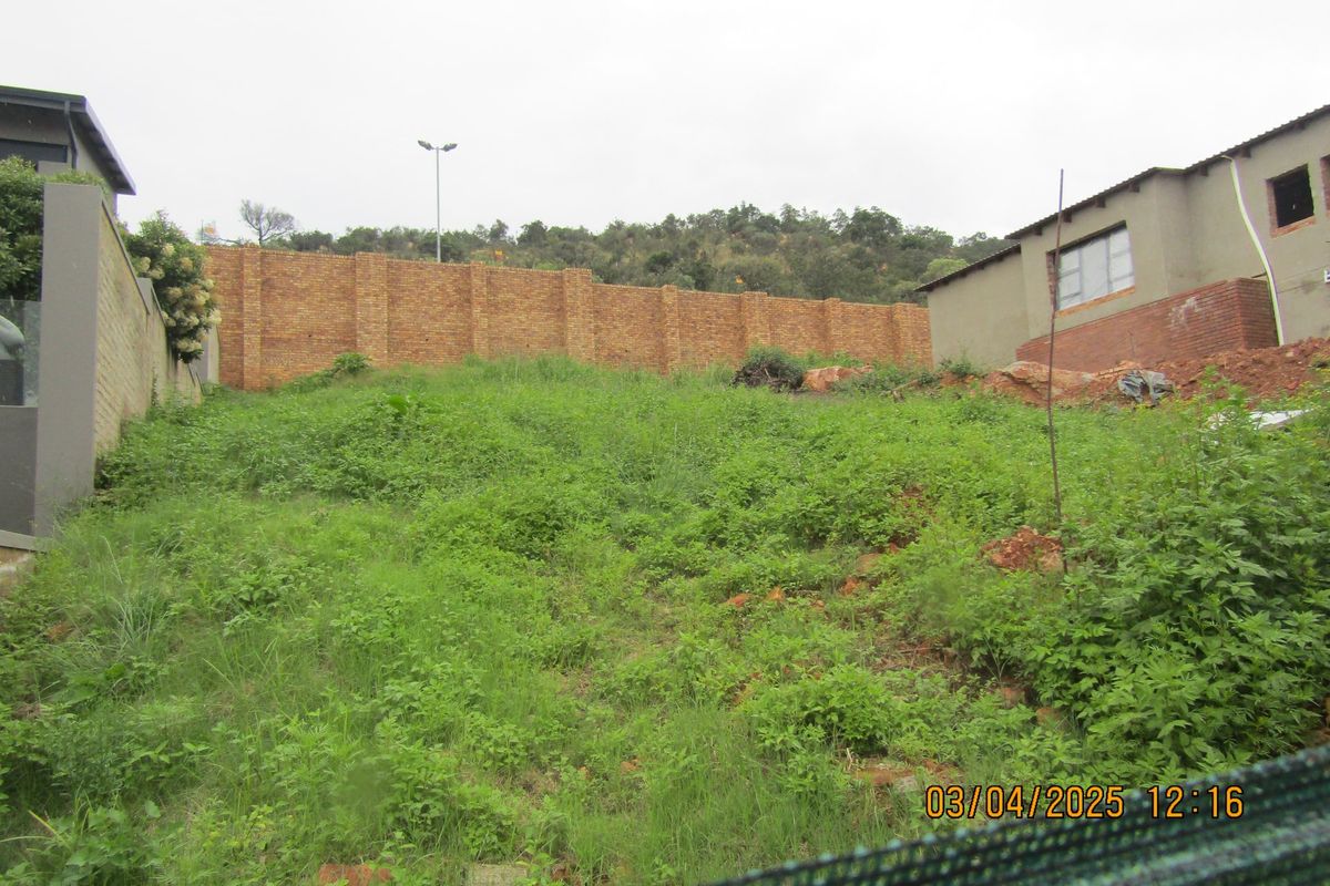 A sloped, undeveloped residential plot featuring a brick boundary wall and overgrown vegetation, situated between existing neighboring properties.