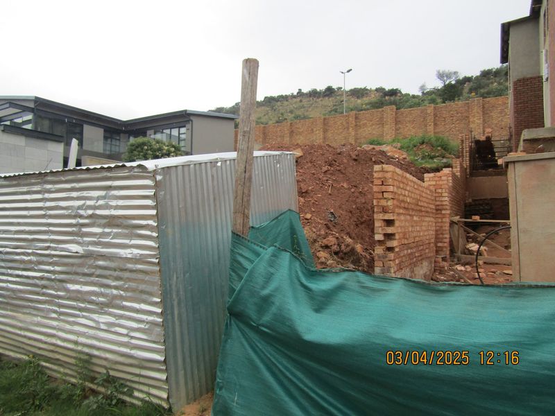 View of a residential plot under construction, featuring unfinished brick retaining walls, a corrugated metal shed, and temporary site fencing.