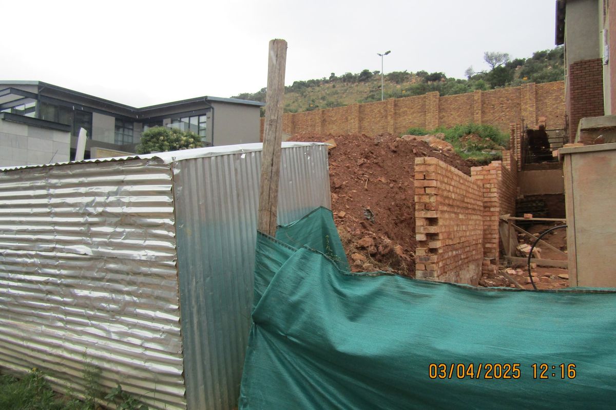 View of a residential plot under construction, featuring unfinished brick retaining walls, a corrugated metal shed, and temporary site fencing.