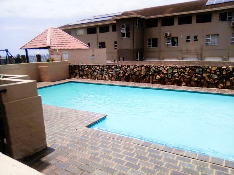 A communal swimming pool area featuring a paved deck, stone retaining wall, and a small thatched gazebo structure.