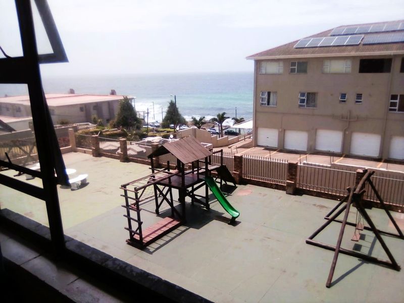 Elevated view overlooking a communal courtyard with playground equipment, perimeter fencing, and a coastal backdrop.