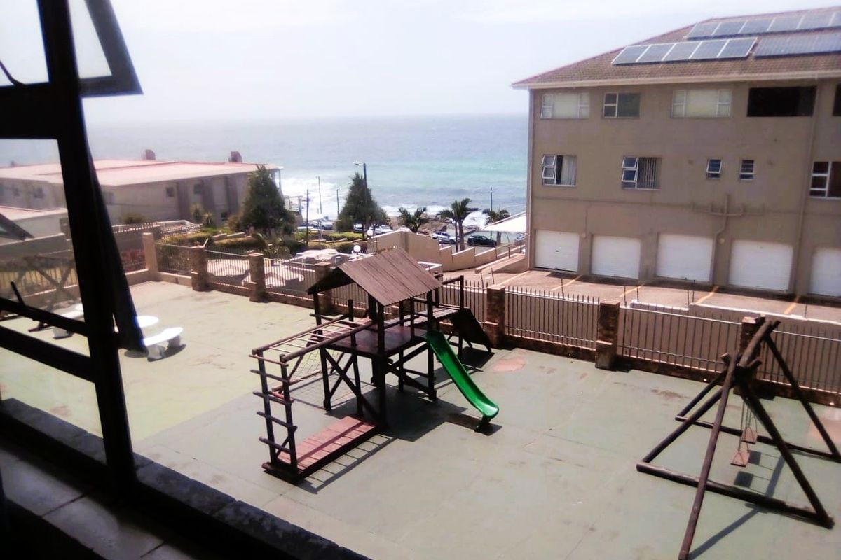 An elevated view of a communal courtyard featuring a playground area, perimeter fencing, and a view of the nearby ocean.