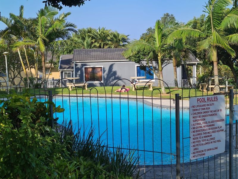 A well-maintained communal swimming pool area featuring paved surrounds, established palm trees, and secure metal fencing within a townhouse complex.