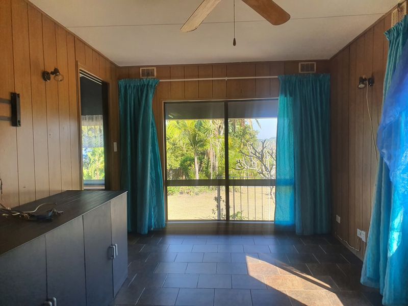 A living room featuring tiled flooring, wood-look wall paneling, a ceiling fan, and a large sliding glass door providing access to the garden.