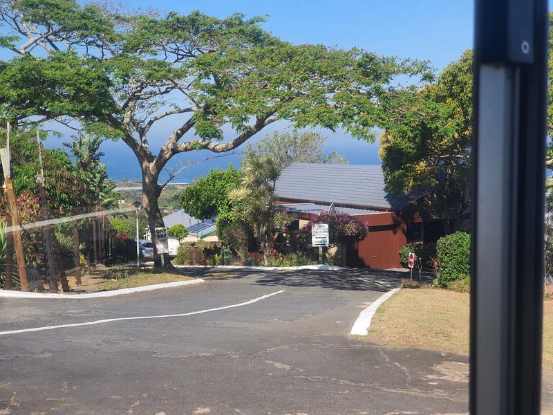 A street-level view of the townhouse complex entrance, featuring mature landscaping, paved access roads, and a view of the distant coastline.