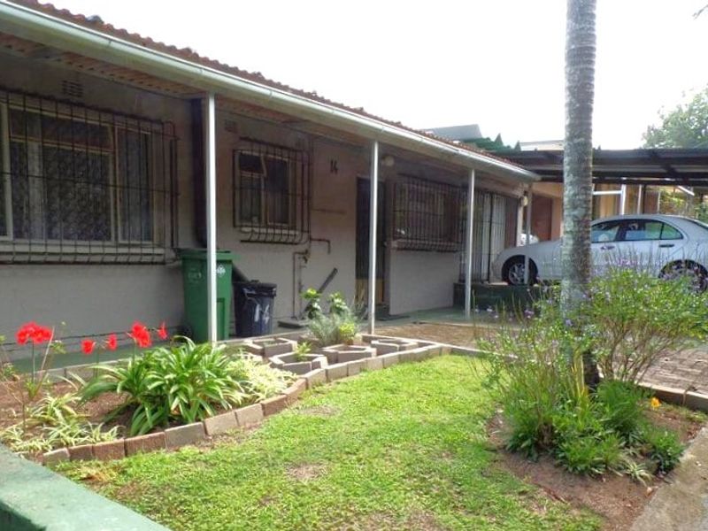 Front elevation of a townhouse featuring a covered patio, security bars on windows, and a small garden area with a paved driveway and carport.