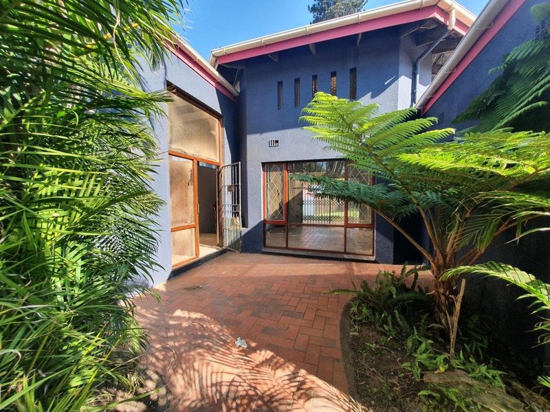 A rear courtyard featuring brick paving, established garden greenery, and access to the home through sliding glass doors and a security gate.