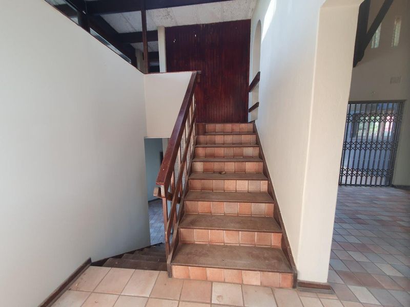 A spacious hallway featuring a tiled staircase with a wooden handrail, exposed timber ceiling beams, and a security gate leading to an adjacent area.