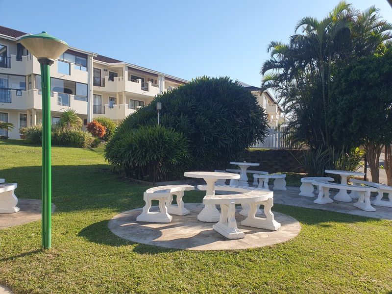 A communal garden area featuring fixed concrete seating and tables set within a landscaped lawn, adjacent to the apartment building complex.