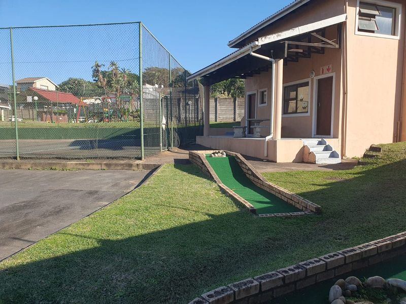 Outdoor recreational area featuring a fenced tennis court and a mini golf course adjacent to a covered patio and building exterior.