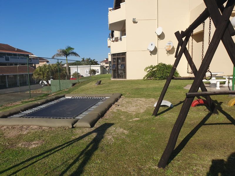 Rear exterior view of the apartment building featuring a communal lawn area with recreational equipment and perimeter fencing.