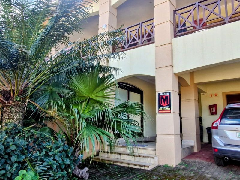 Front elevation of a townhouse featuring a covered entrance, upper-level balcony with wooden railings, and a paved driveway area.