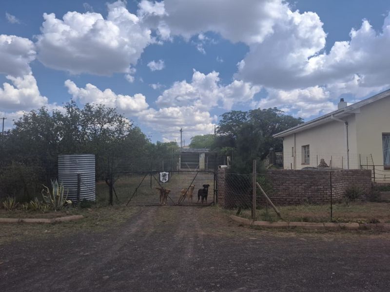 Front view of the property featuring a gravel driveway, metal gate, brick boundary wall, and a water tank in a rural setting.