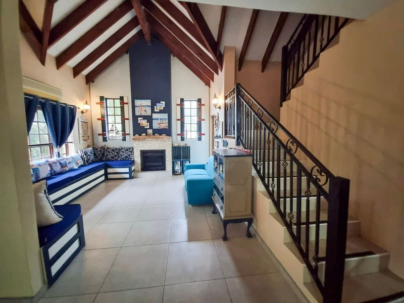 Spacious living room featuring a vaulted ceiling with exposed timber beams, a central fireplace, tiled flooring, and a decorative wrought iron staircase.