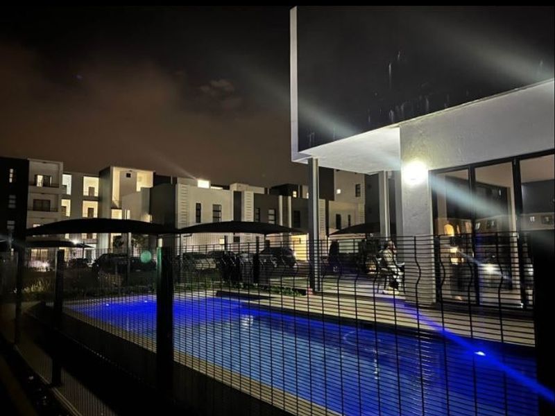 Night view of a modern apartment complex featuring a swimming pool, secure metal fencing, and illuminated outdoor patio area.
