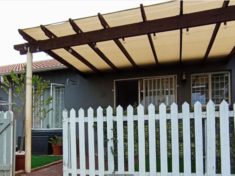 A private outdoor patio area featuring a timber pergola with shade cloth, enclosed by a white picket fence and secure access gate.