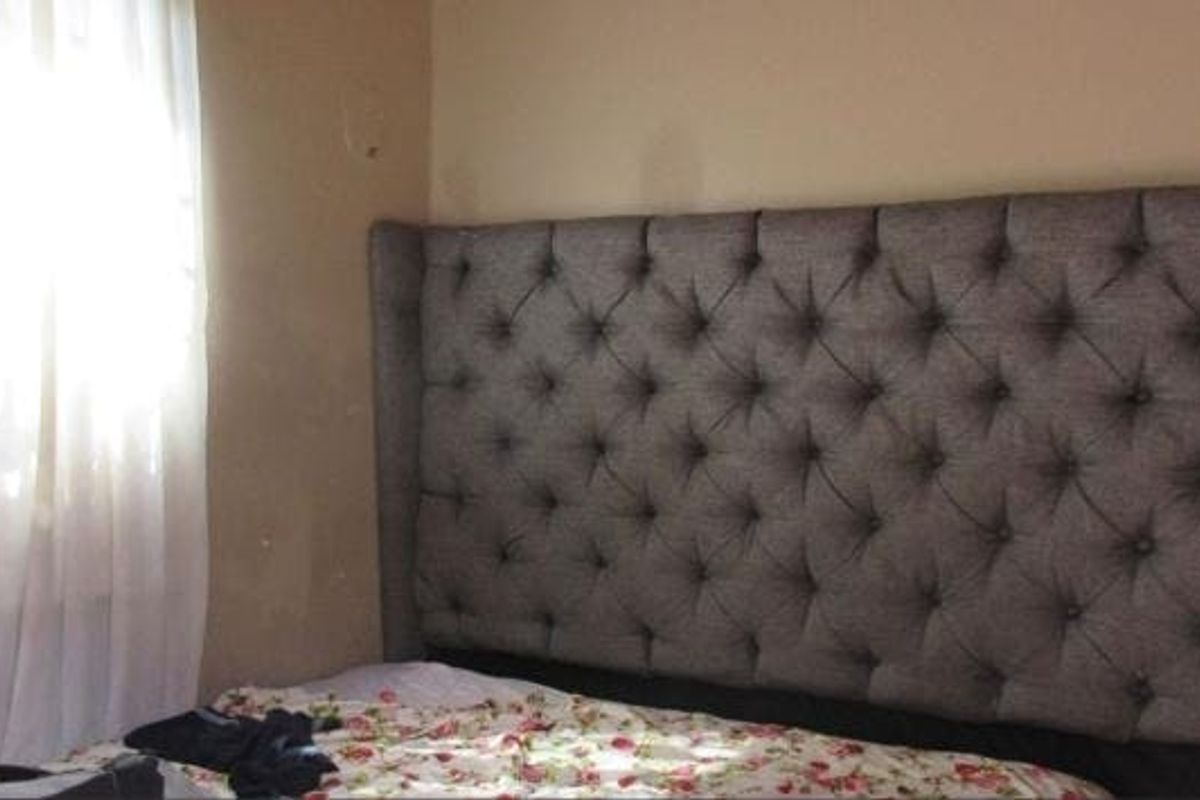 A bedroom interior featuring a tufted headboard and a window with curtains, showing signs of wear on the wall surfaces.