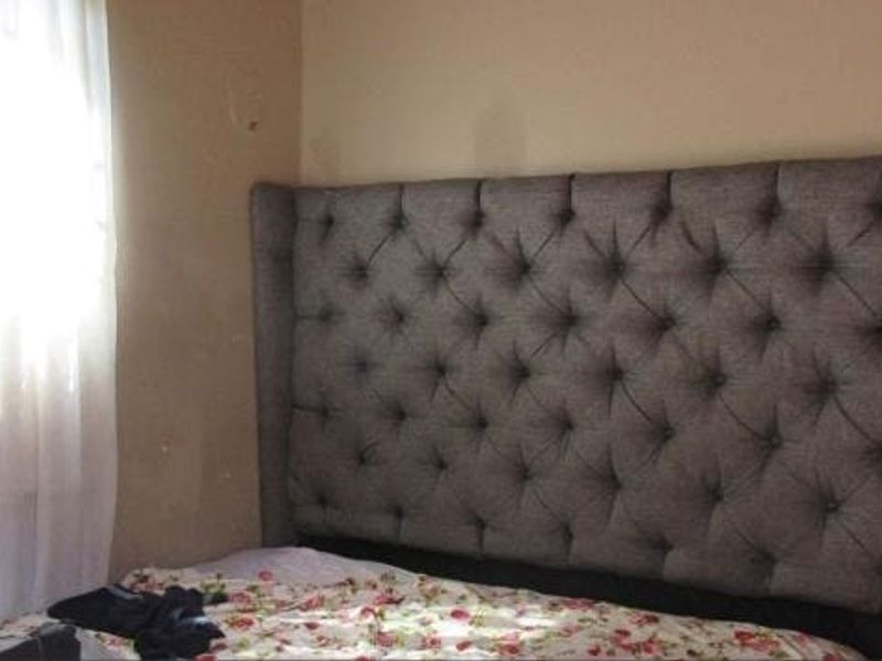 A bedroom interior featuring a tufted headboard and a window with curtains, showing signs of wear on the wall surfaces.