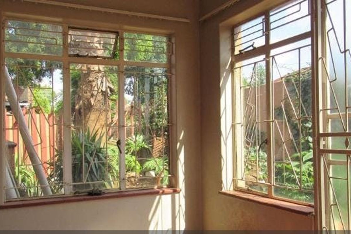 An empty living area featuring large steel-framed windows with security bars, offering views of the garden and natural light.