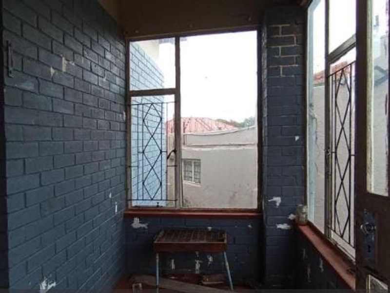 An enclosed balcony or sunroom featuring painted brick walls, large windows with security bars, and a tiled floor, currently in need of cosmetic refurbishment.