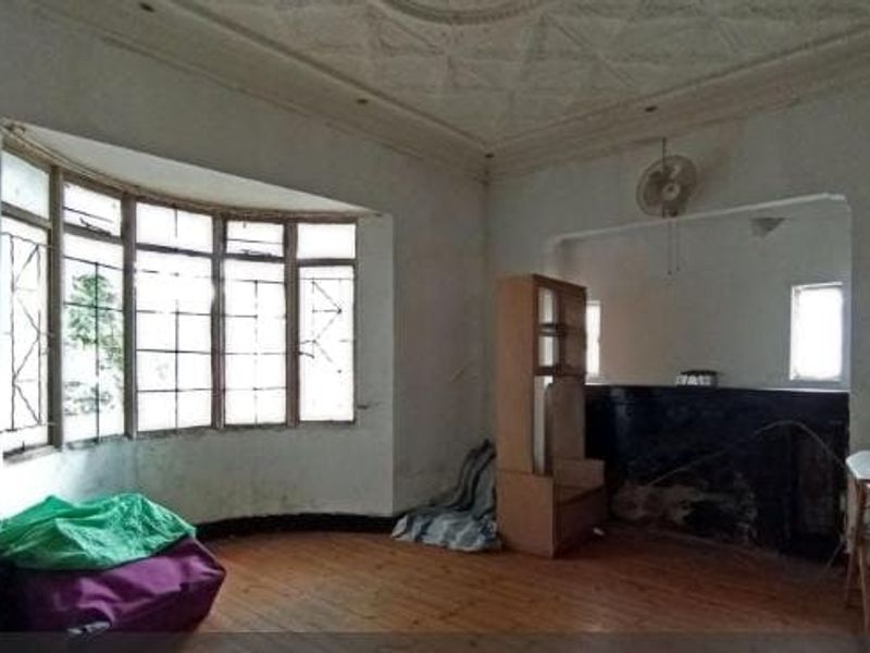 Spacious living room featuring a large bay window, ornate ceiling detailing, and original hardwood flooring, currently in need of significant renovation and cosmetic repair.