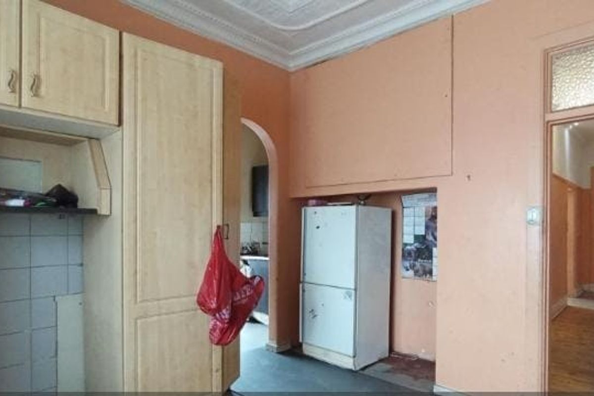 A dated kitchen area featuring built-in cabinetry, tiled splashbacks, and an ornate ceiling cornice, leading into a hallway with original wooden flooring.