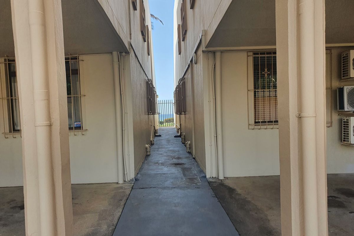 A narrow exterior walkway between apartment building blocks, featuring concrete flooring, security-barred windows, and wall-mounted air conditioning units.