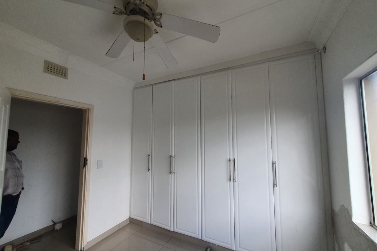 A bedroom featuring built-in cupboards, a ceiling fan, and tiled flooring, with some visible wall plaster damage near the window.