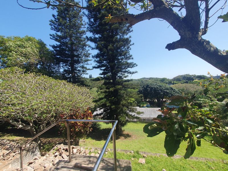 A scenic elevated view overlooking a lush green garden with mature trees, featuring a stone retaining wall and stainless steel handrail.