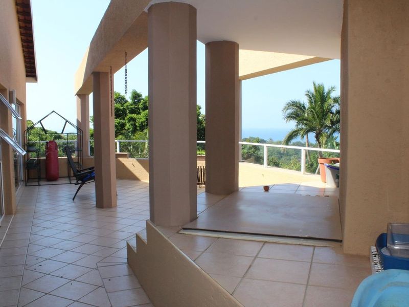 Spacious covered patio featuring tiled flooring, structural columns, and a glass balustrade, offering scenic views of the surrounding landscape and ocean.
