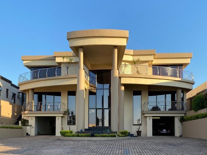 Modern double-story residence featuring a grand entrance portico, expansive glass facade, dual balconies with stainless steel railings, and a spacious paved driveway.