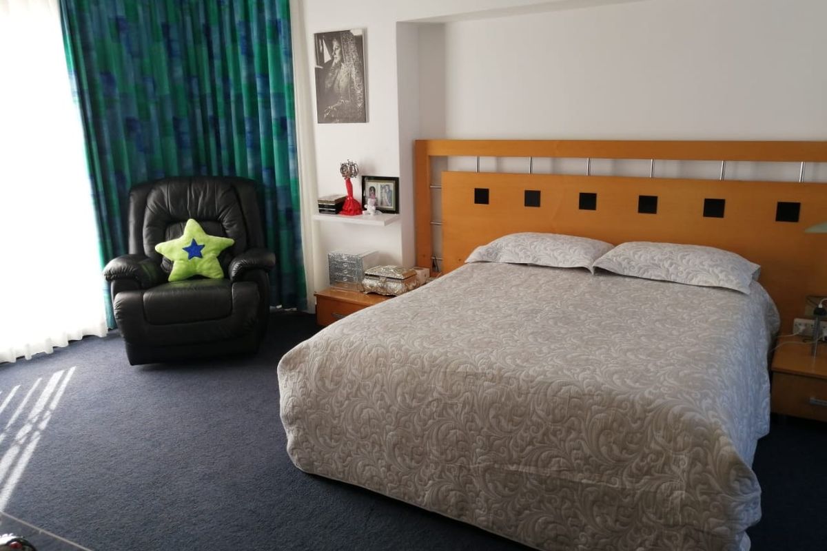 A bedroom featuring a built-in wooden headboard and dark carpeting, with access to natural light through large window curtains.