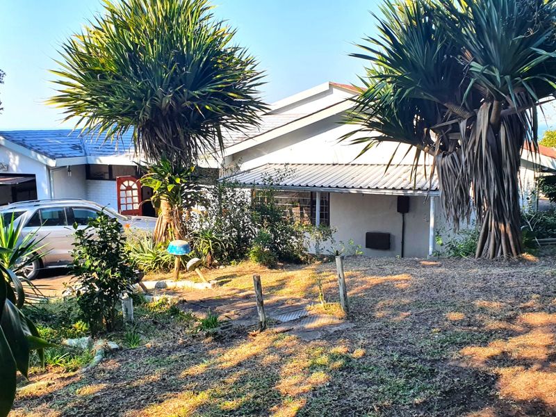 Front view of a single-story residential house featuring a mix of tiled and metal roofing, set within a large, established garden area.