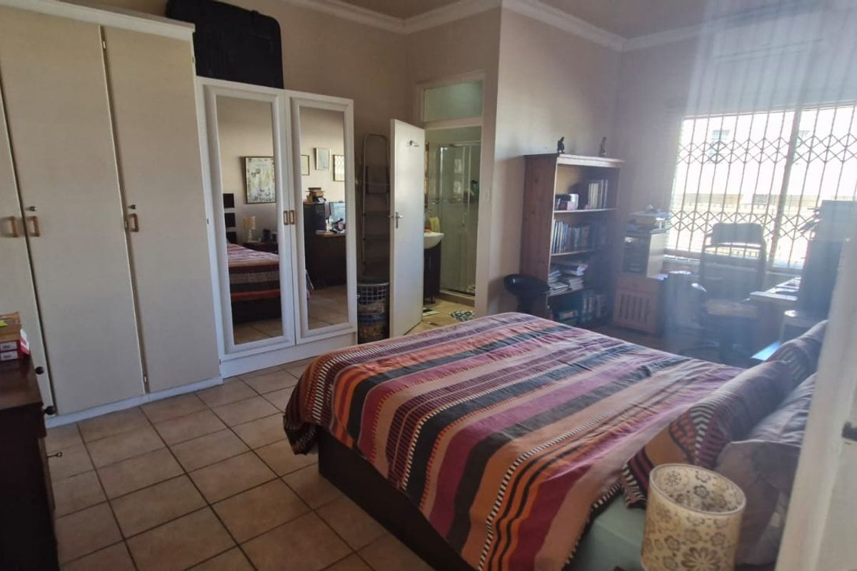 A functional bedroom featuring built-in cupboards, tiled flooring, and an en-suite bathroom, with natural light entering through a window with security bars.