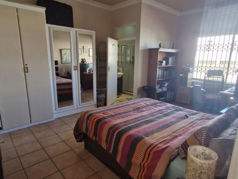 A functional bedroom featuring built-in cupboards, tiled flooring, and an en-suite bathroom, with natural light entering through a window with security bars.