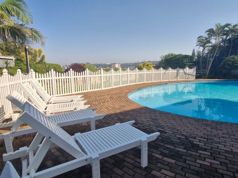 A communal swimming pool area featuring brick paving and a white picket fence, set within a landscaped residential complex.